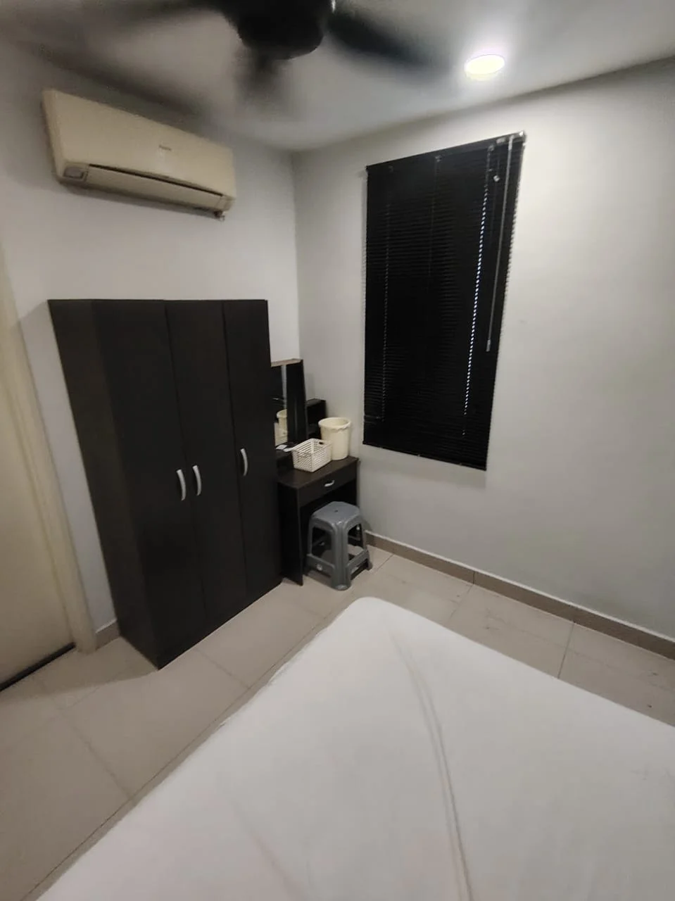 Bedroom features: white painted walls, tile flooring, ceiling fan, 1 wall-mounted split AC, blackout blinds, dark wood wardrobe, dark wood bedside table, mirror on table, small stool.